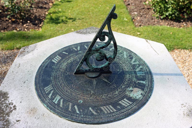 Sundial on stone plinth. Metal sundial on stone plinth with gnomon sharply in focus with background of flower beds and lawn in full sun with the time clearly stock images