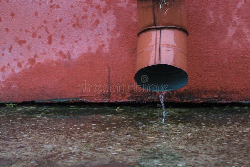 Trickle of water flowing from red metal drain pipe on cement on the blind area of the building with red facade. A thin trickle of water flowing from the red royalty free stock image