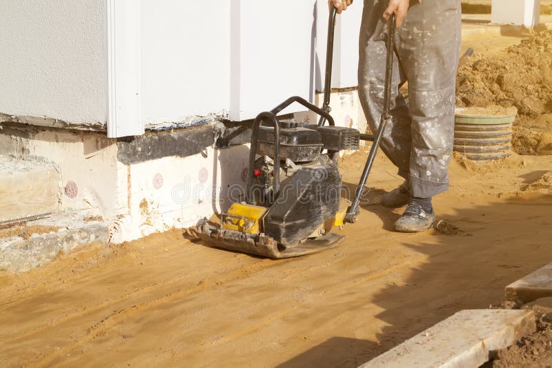 Worker compresses sand in blind area around building with special working tool & x28;tamping machine stock photos