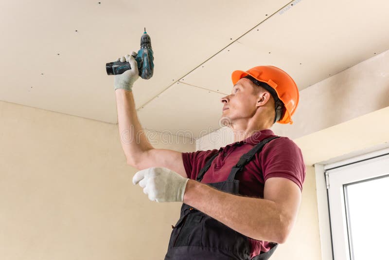 Worker is using screws and a screwdriver to attach plasterboard to the ceiling. Installation of drywall. Worker is using screws and a screwdriver to attach stock photos